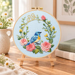 Blue bird inside pink rose wreath embroidery kit finished hoop displayed on a wooden stand as cosy tabletop decor