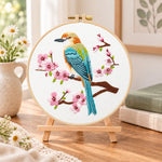 Colourful bird amid pink cherry blossoms embroidery kit finished hoop displayed on a wooden stand as cosy tabletop decor