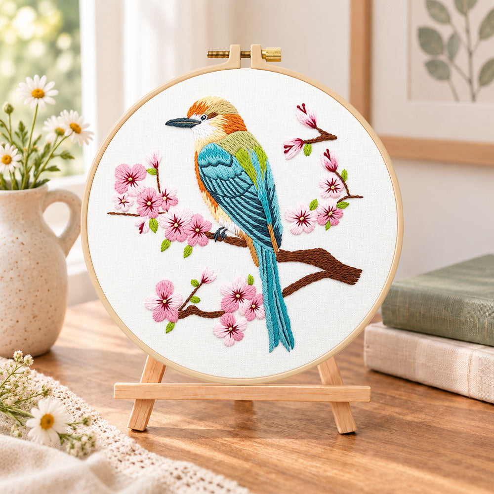 Colourful bird amid pink cherry blossoms embroidery kit finished hoop displayed on a wooden stand as cosy tabletop decor