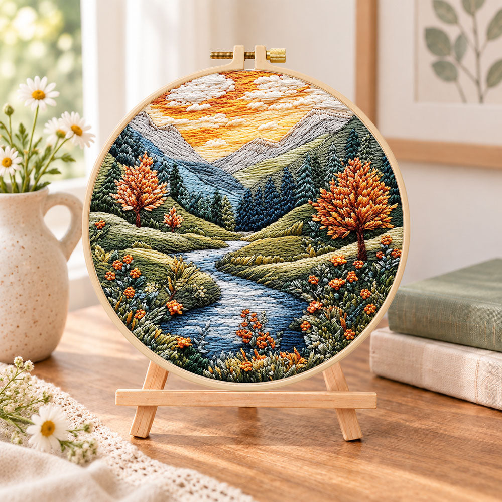 Golden hour mountain river embroidery kit finished hoop displayed on a wooden stand as cozy tabletop decor