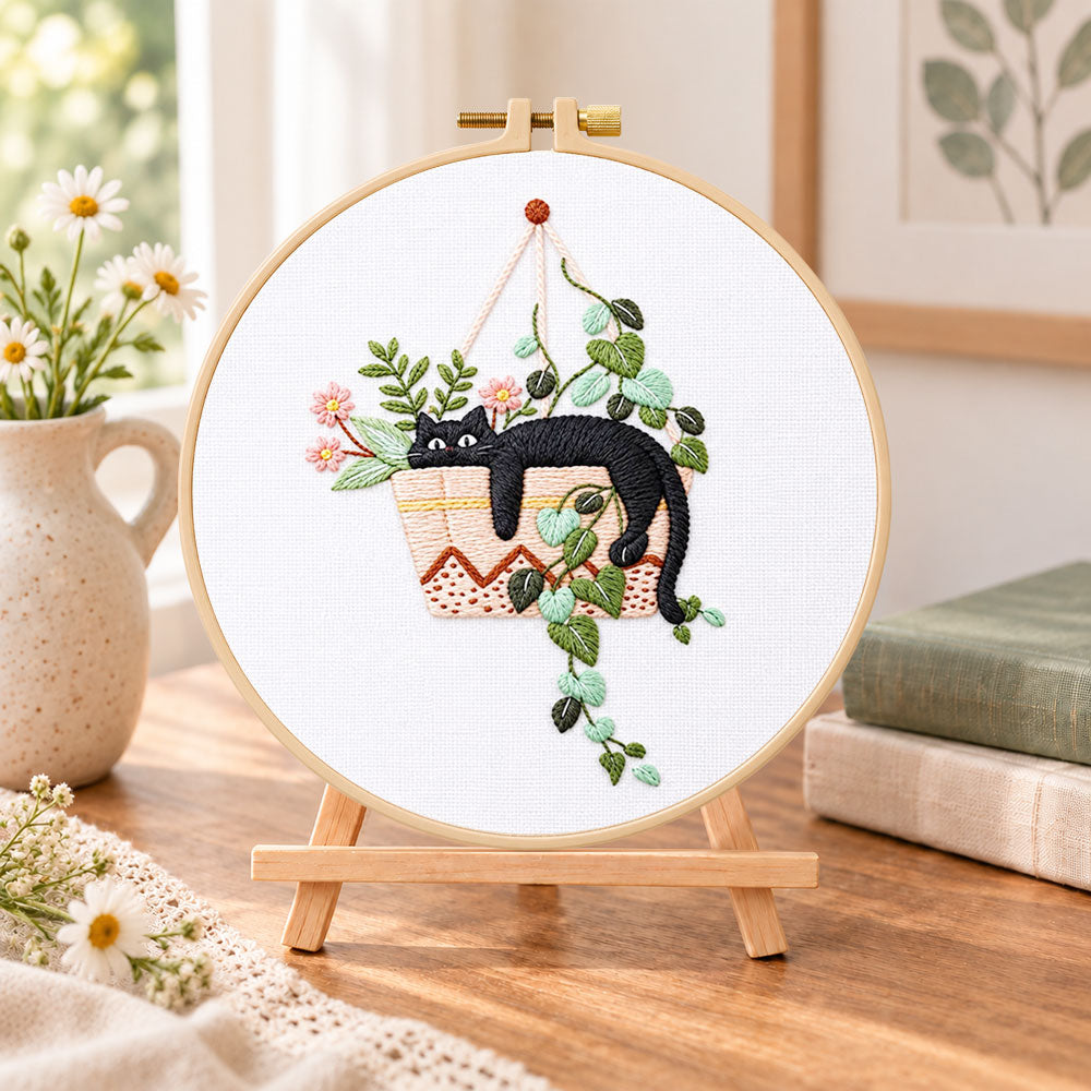 Lazy cat in hanging planter embroidery kit finished hoop displayed on a wooden stand as cosy tabletop decor