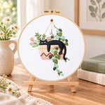 Lazy cat in hanging planter embroidery kit finished hoop displayed on a wooden stand as cosy tabletop decor