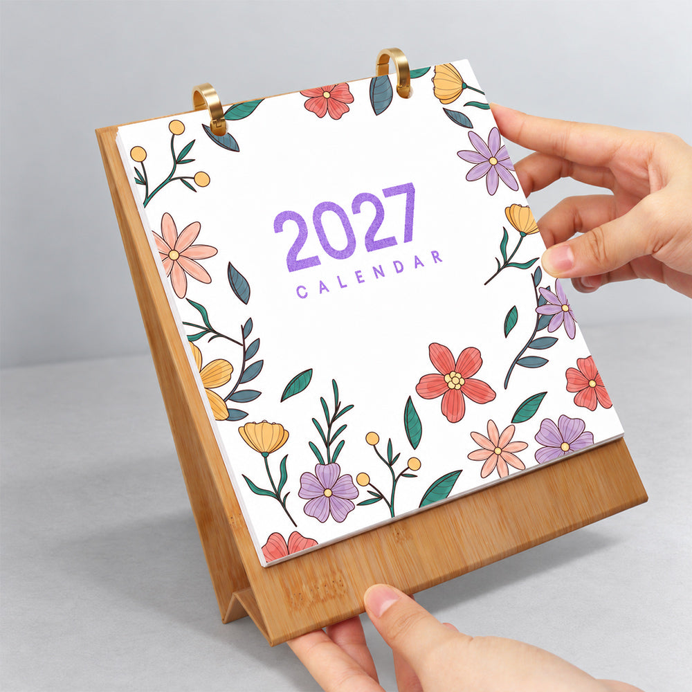 Hands flipping the floral cover page on a wooden desk calendar with gold metal binder rings