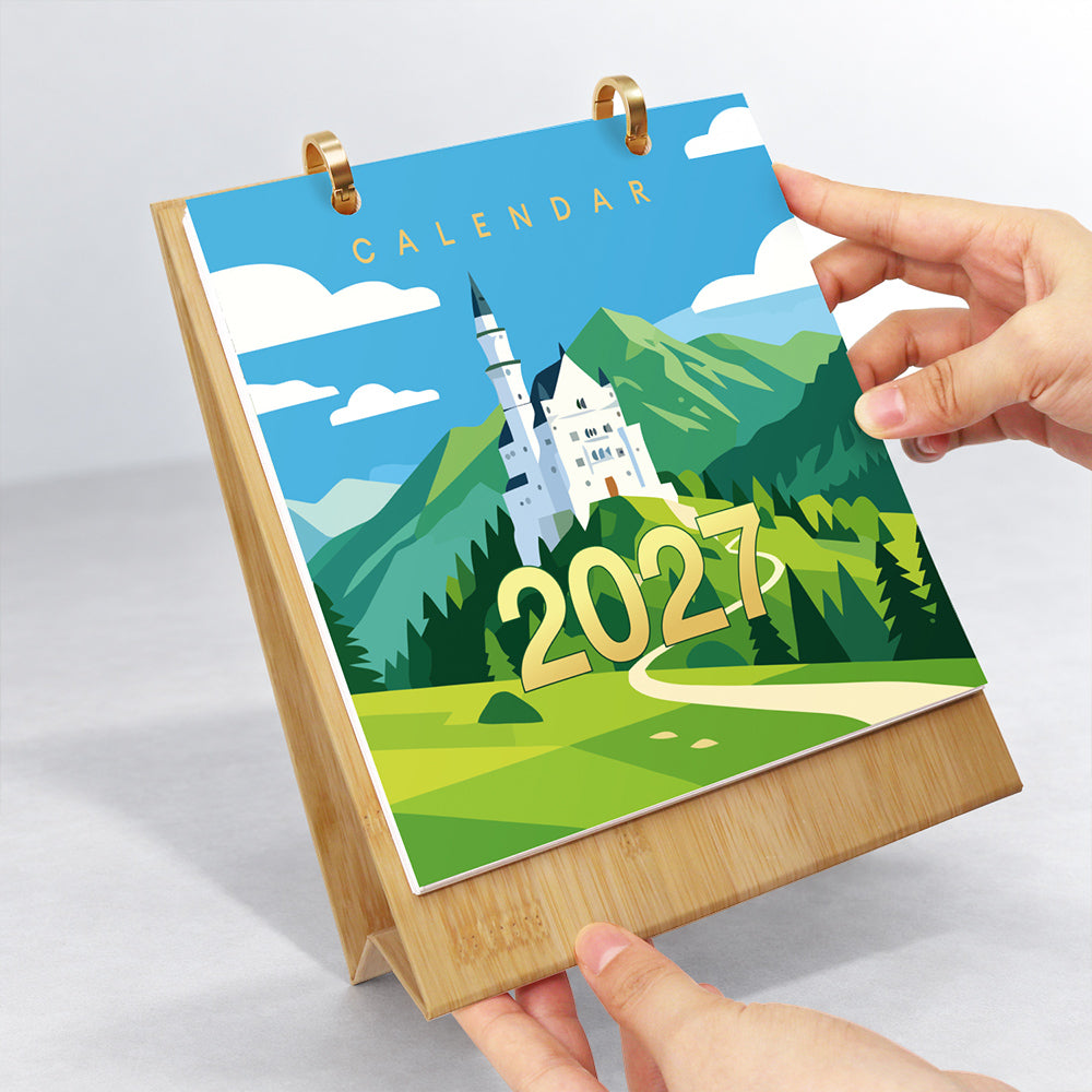 Hands flipping the castle-themed cover page on a wooden desk calendar with gold metal binder rings