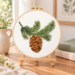 Pine branch and cone embroidery kit finished hoop displayed on a wooden stand as cozy tabletop decor