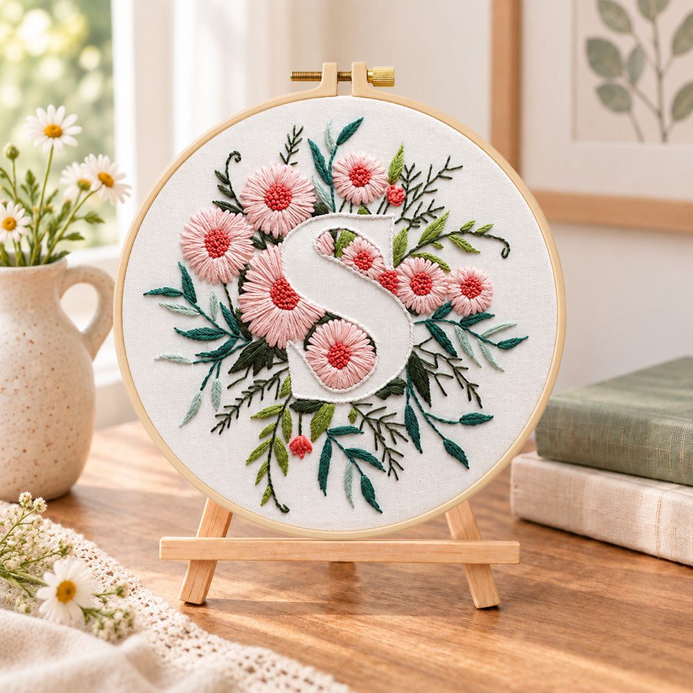 Pink daisy blooms and letter s embroidery kit finished hoop displayed on a wooden stand as cosy tabletop decor