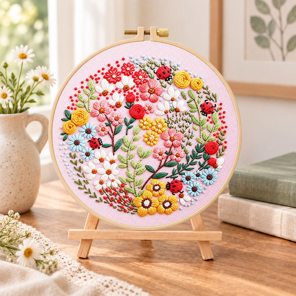 Pink floral art with ladybugs embroidery kit finished hoop displayed on a wooden stand as cosy tabletop decor