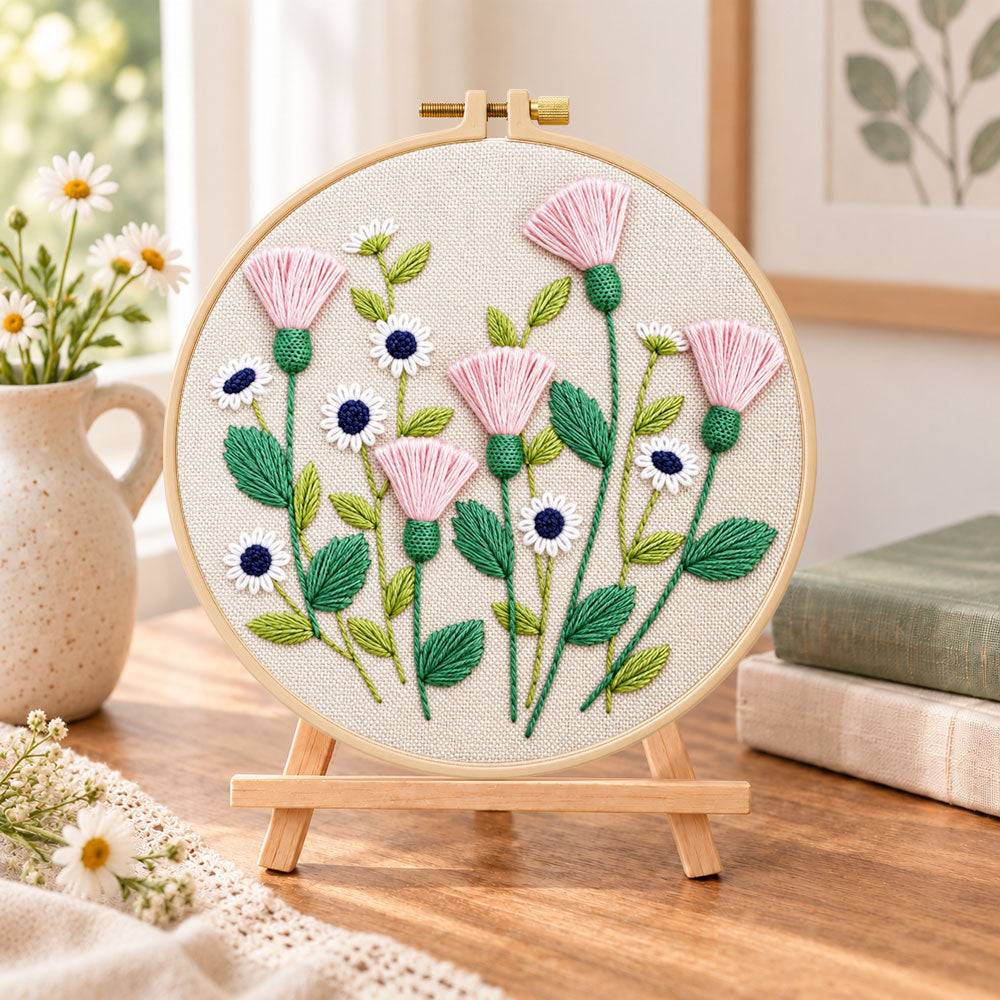 Pink thistle and daisies embroidery kit finished hoop displayed on a wooden stand as cosy tabletop decor