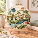Tropical riverside hamlet embroidery kit finished hoop displayed on a wooden stand as cosy tabletop decor