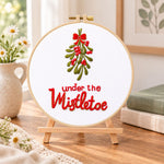 Under the mistletoe embroidery kit finished hoop displayed on a wooden stand as cozy tabletop decor