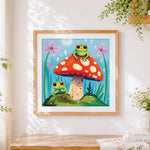 Finished whimsical frogs on a toadstool cross stitch artwork displayed